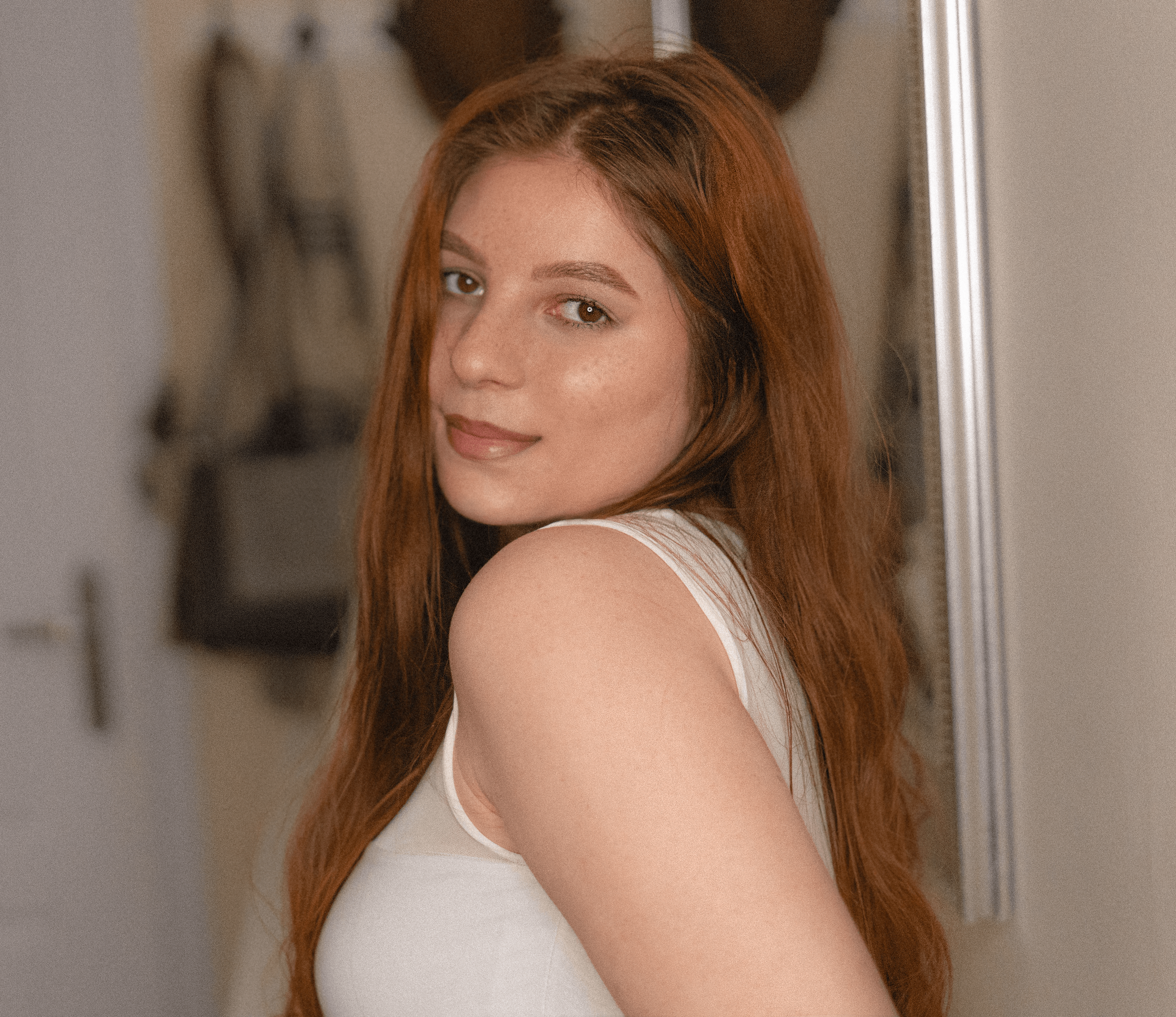 Woman with long red hair wearing a white sleeveless top in a room.